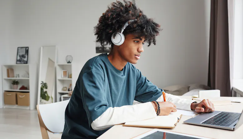 A young man sits at a desk in a bedroom taking notes from his laptop computer.