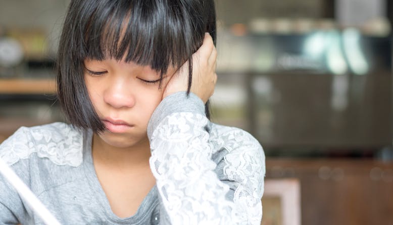 A young girl sits at a table doing homework looking frustrated.