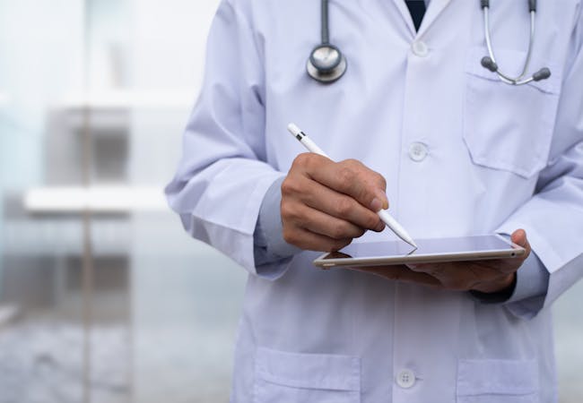 A doctor is shown from the shoulders to the waist holding a tablet with a pen.