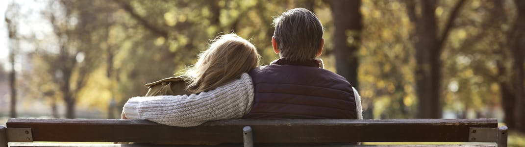 An elderly couple on a bench in a park outside sit and embrace.
