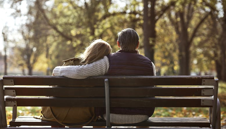 An elderly couple on a bench in a park outside sit and embrace.