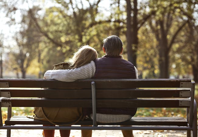 An elderly couple on a bench in a park outside sit and embrace.
