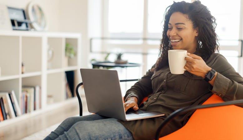 A young black woman sits looking at her laptop and smiling, holding a mug in her hand.
