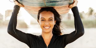 A woman smiling holds a surf board over her head.