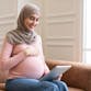 A pregnant woman wearing a headscarf, sitting on a couch and looking at a tablet.
