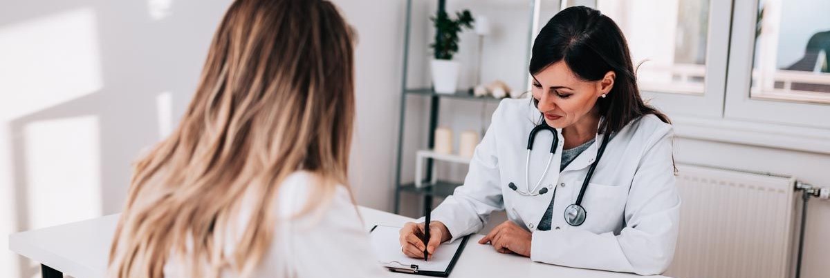 A female doctor writes notes while interacting with a female patient in an office.