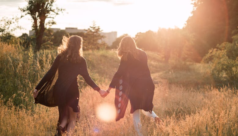 Two women hold hands while walking through a meadow. 