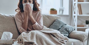 A woman on the couch with the flu blows her nose into a tissue. 