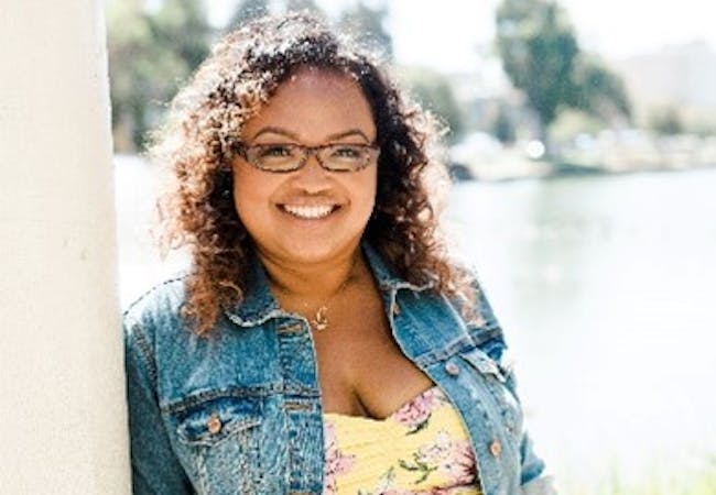 Brenda Kong in a floral jumpsuit and jean jacket, smiling and leaning against a column outside.