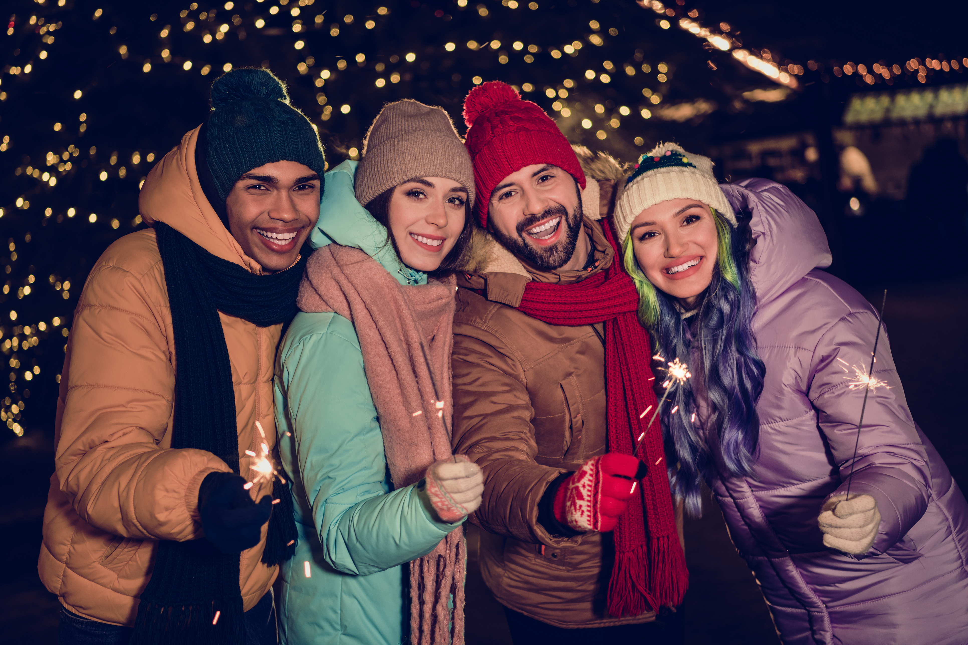 Friends gather outside with sparklers and holiday lights behind them.