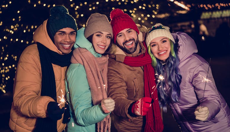 Friends gather outside with sparklers and holiday lights behind them.