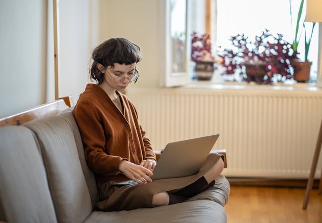 A woman sits on a couch in an apartment and looks at a laptop on her lap.