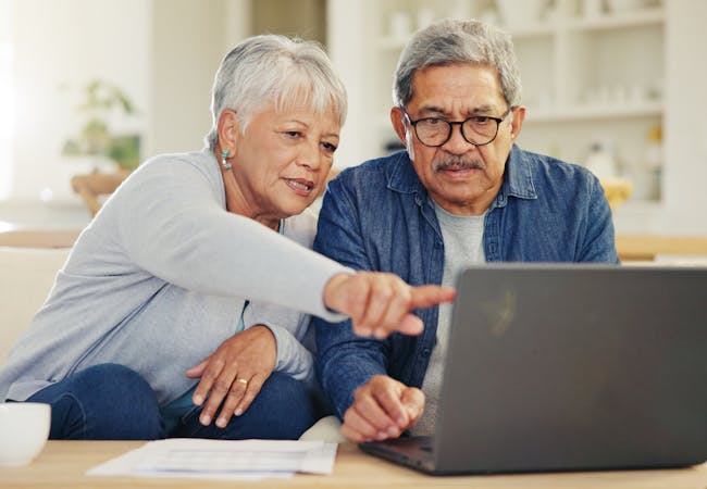 An older Latine couple looks at a laptop together while sitting on a couch.