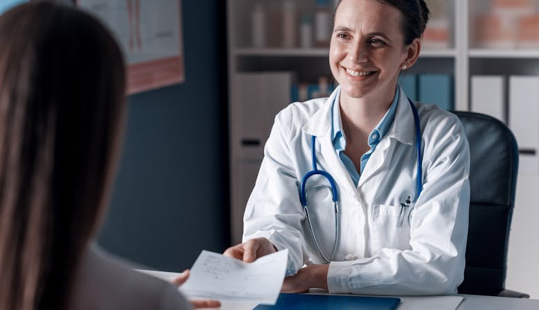 A woman doctor hands a prescription to a patient across a desk.
