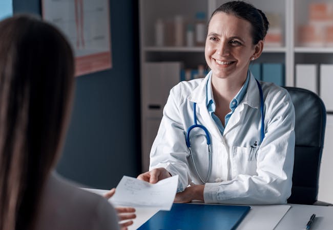 A woman doctor hands a prescription to a patient across a desk.