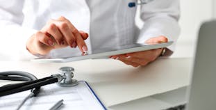 A doctor points at a tablet in their hands, a laptop, paperwork, and a stethoscope are in the foreground.
