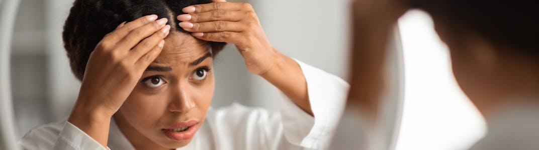 A black woman looks at her scalp in the mirror while wearing a bathrobe.