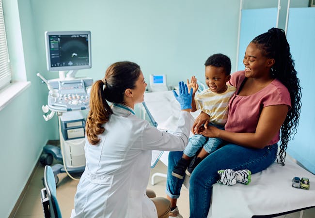 A Black mother and son sit in a doctor's office. A doctor high-fives the son,