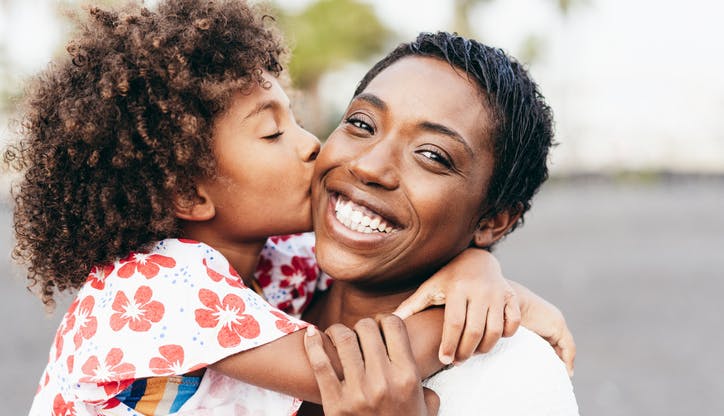 A young Black girl kisses her smiling mom on the cheek.