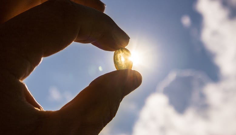 Vitamin D supplement held up in a hand with the sun shining behind it.