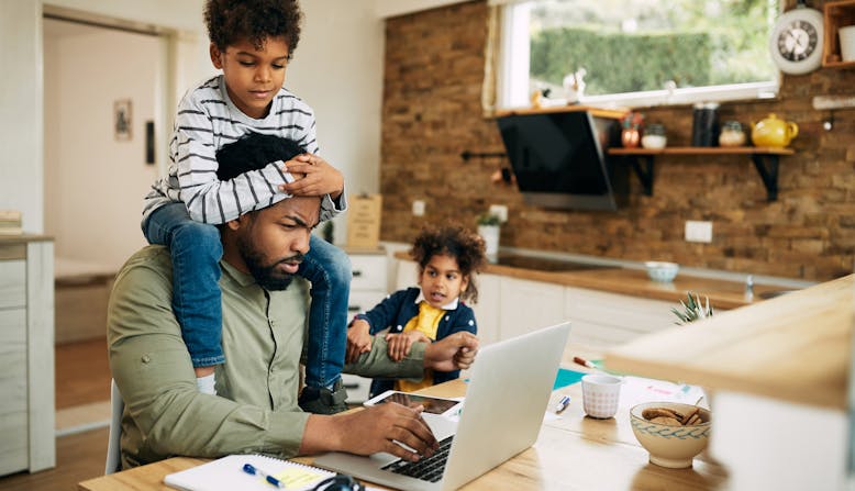 A father works from home with a child crawling on him and another in the background.