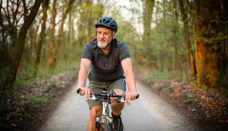 Middle aged to older man with grey beard riding a mountain bike on a paved trail through a wooded area.