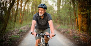 Middle aged to older man with grey beard riding a mountain bike on a paved trail through a wooded area.