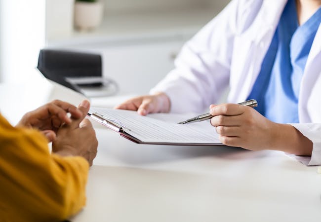 A doctor holds a clipboard and talks to a patient at a desk.
