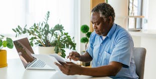 An elderly man looks at documents while sitting at a desk with a laptop.