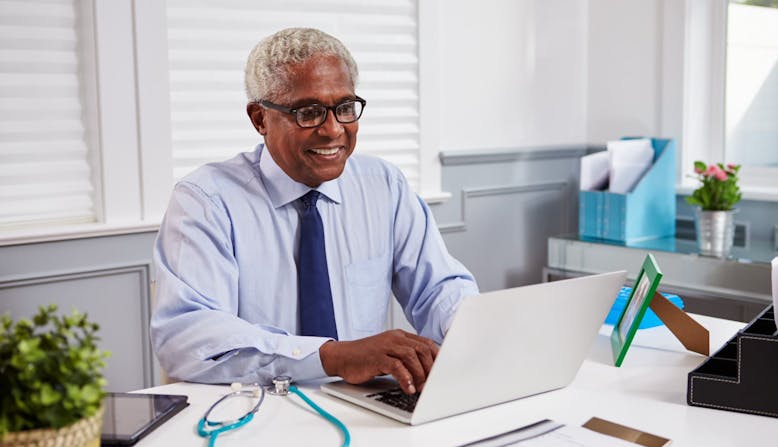 A male medical professional sitting at his desk, smiling and working on a laptop. 