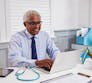 A male medical professional sitting at his desk, smiling and working on a laptop.