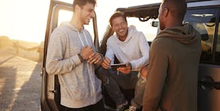 Three men near a car, one sitting in the driver seat with the door open.