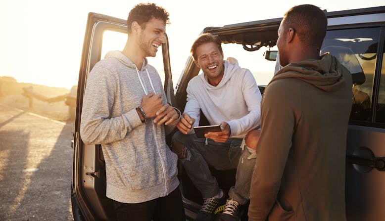 Three men near a car, one sitting in the driver seat with the door open.