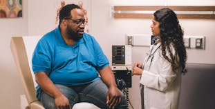A larger bodied man sits in a doctor's office with a woman doctor standing next to him.