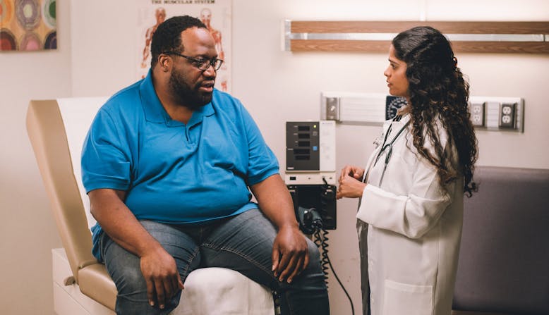 A larger bodied man sits in a doctor's office with a woman doctor standing next to him.
