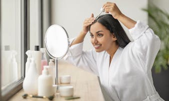 A woman looks in a mirror and applies a product to her scalp.