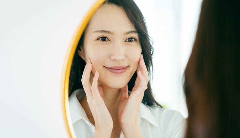 A woman looks in the mirror smiling and touching her face.