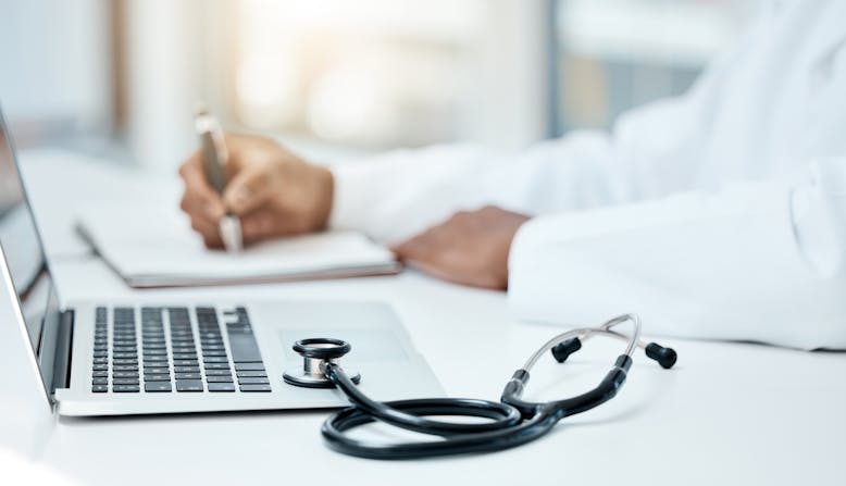 A doctor writing notes and looking at a laptop computer.