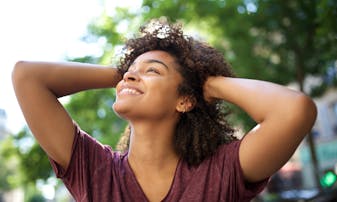 A woman smiles and touches her hair, standing outside.