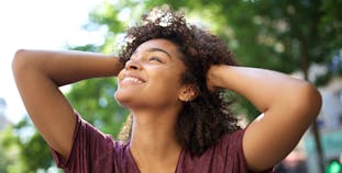 A woman smiles and touches her hair, standing outside.