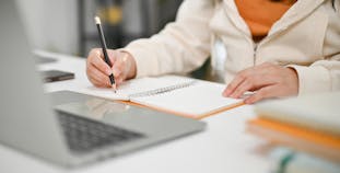 A woman writes notes in a notebook while looking at a laptop computer.