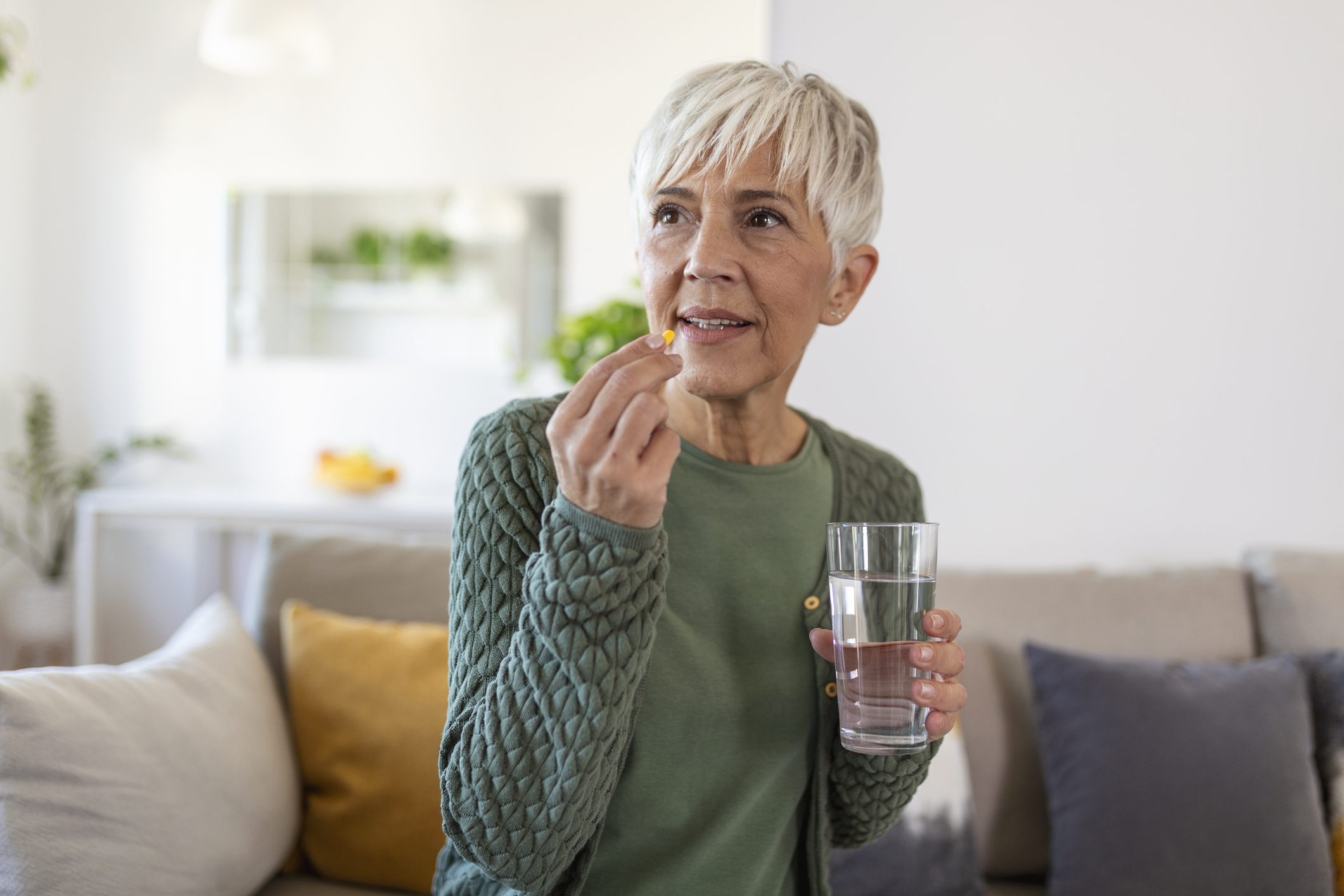 An elderly woman is sitting on a couch taking a pill with a glass of water.