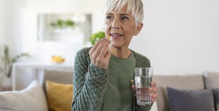 An elderly woman is sitting on a couch taking a pill with a glass of water.