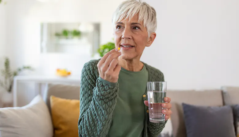 An elderly woman is sitting on a couch taking a pill with a glass of water.