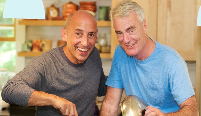 Two older men smiling in a kitchen.