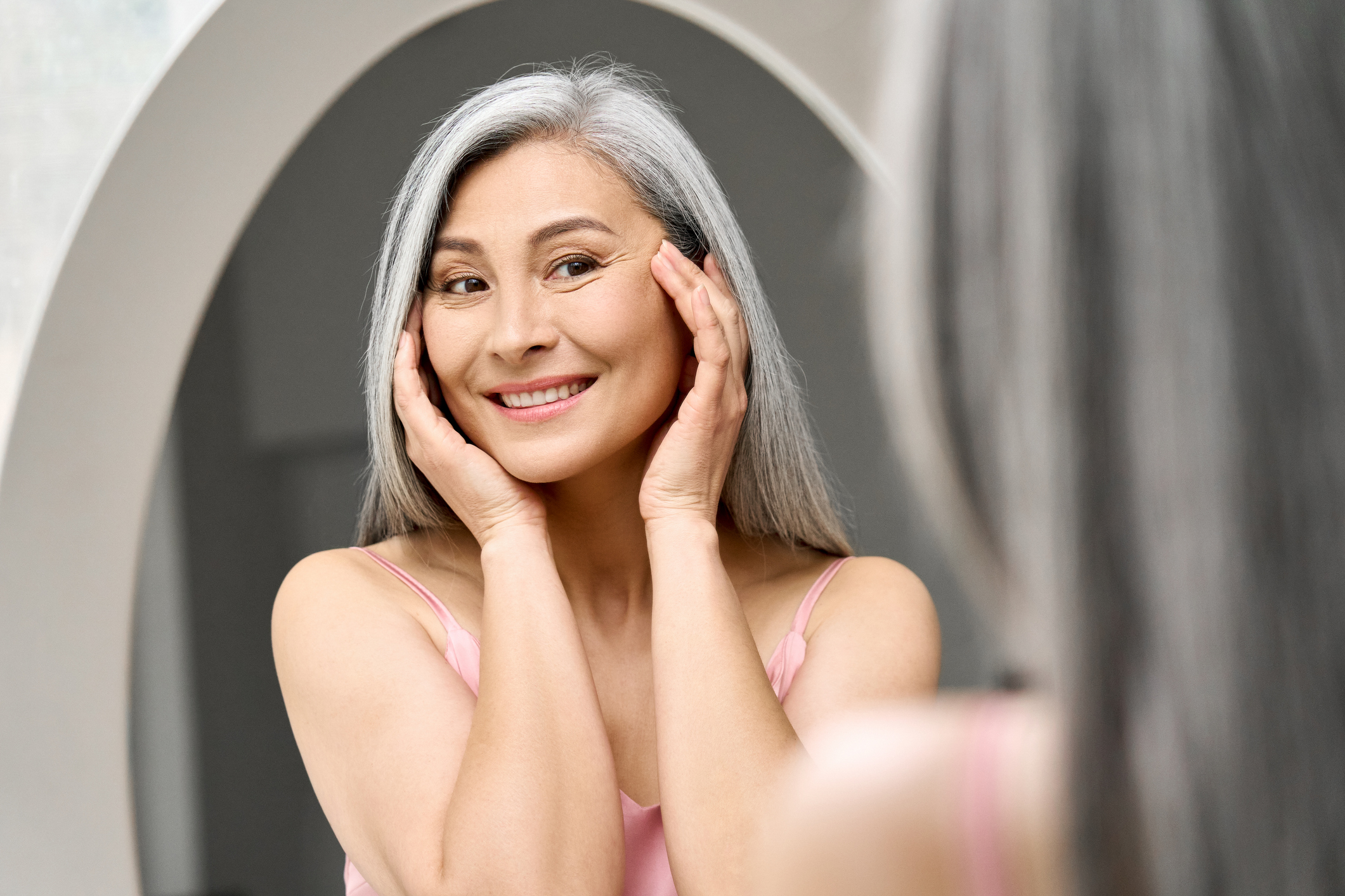 An elderly woman looks at her skin in the mirror.