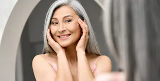 An elderly woman looks at her skin in the mirror.