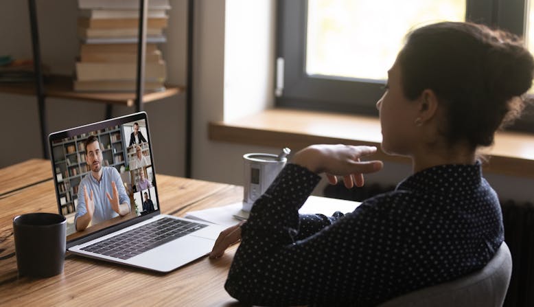 Woman sitting at a desk in front of a laptop with images of several people, the main one a man gesturing with his hands.