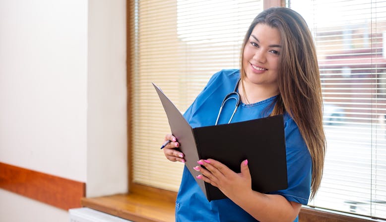 A smiling doctor standing in a hospital hallway next to a window, wearing a blue uniform and stethoscope, and holding a folder.