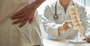 A person holding their back in pain while a doctor points to a model of the spine.